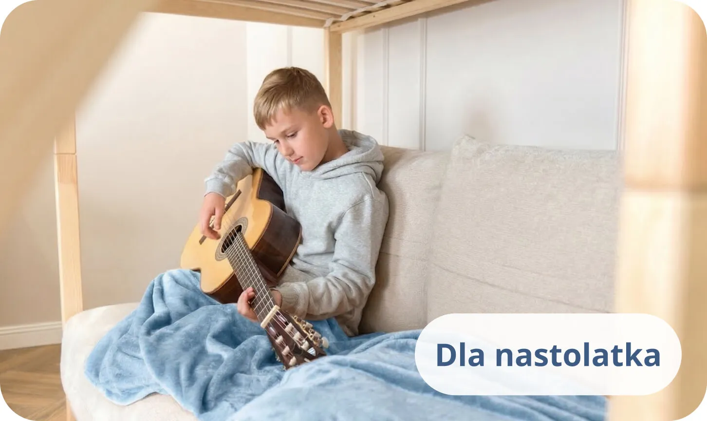 Nastolatek grający na gitarze akustycznej na łóżku przykryty niebieskim kocem.